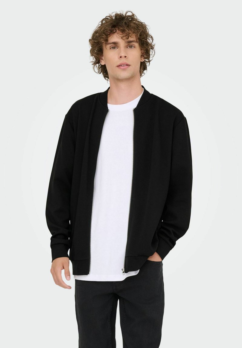 Sort zip-up bomberjakke med ribbede cuff og krave, iført over en hvid T-shirt, parret med mørke jeans. Ensartet baggrund.