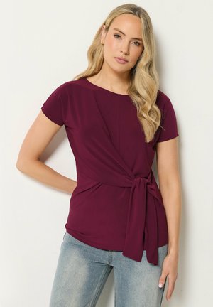 Lyshåret kvinde iført en kortærmet bordeaux top med bindebånd i taljen og lyseblå jeans, stående mod en ensfarvet hvid baggrund.