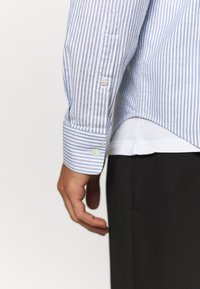 Nahaufnahme eines Arms einer Person, die eine blau-weiß gestreifte, knöpfbare Hemdmanschette trägt, schwarze Hose und weißes Unterhemd sind sichtbar.