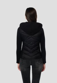 Črna jakna s kapuco s prešito, napihnjeno hrbtno stranjo v vzorcu diamantov, kombinirana s temnimi kavbojkami. Gladek teksturni material s prilagojeno zasnovo.