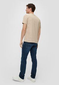 Bež t-shirt od pamuka s kratkim rukavima, u kombinaciji s tamnoplavim trapericama i bijelim tenisicama, prikazan sa stražnje strane.