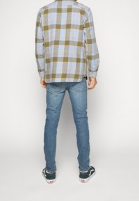 Camicia a scacchi in tonalità verde, blu e giallo, abbinata a jeans aderenti azzurri. Le scarpe sono sneakers bianche e nere con lacci.