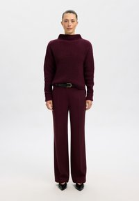 Pull en maille bourgogne avec un col montant associé à un pantalon large de couleur assortie. Une ceinture noire et des chaussures à bout pointu complètent la tenue.