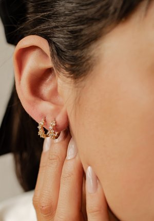 Orecchini a cerchio in oro con accenti di pietre preziose chiare e gialle, caratterizzati da un design floreale, indossati all'orecchio. Pelle liscia e smalto per unghie neutro visibili.