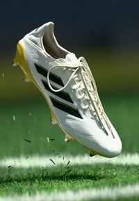 White soccer cleat with black stripes and yellow accents, featuring a smooth upper, lace closure, and visible studs, hovering above grass.