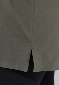 Oliivinvihreä t-paita, jossa on kaareva helma ja sivuhalkiot, valmistettu pehmeästä puuvillamateriaalista. Ominaisuuksiin kuuluvat yksinkertainen ompelu ja sileä pinta.