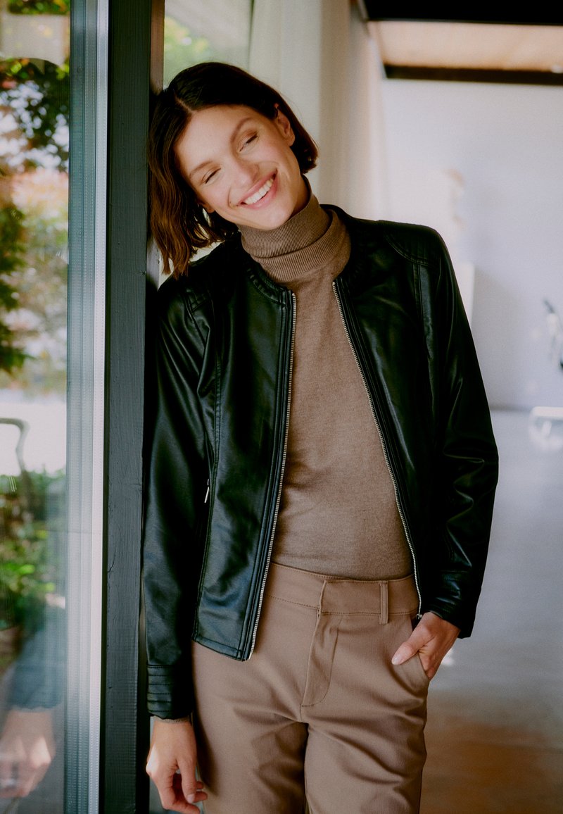 Zwarte leren jas, aansluitende bruine coltrui, beige broek. Het model leunt tegen glas, toont modern ontwerp en kleuren.