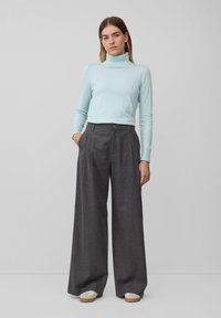Pull à col montant bleu clair associé à un pantalon large à taille haute, gris, avec des plis devant et des poches latérales. Des baskets beiges viennent compléter la tenue.