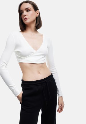 Junge Frau trägt ein weißes, langärmeliges, gekürztes Wickeloberteil und schwarze Hosen mit Kordelzug und posiert mit einer Hand in der Tasche vor einem schlichten Hintergrund.