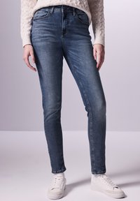 Jeans skinny en denim bleu foncé, avec une taille mi-haute, cinq poches et une texture lisse, associés à des baskets blanches.