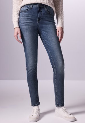 Jeans skinny en denim bleu foncé, avec une taille mi-haute, cinq poches et une texture lisse, associés à des baskets blanches.