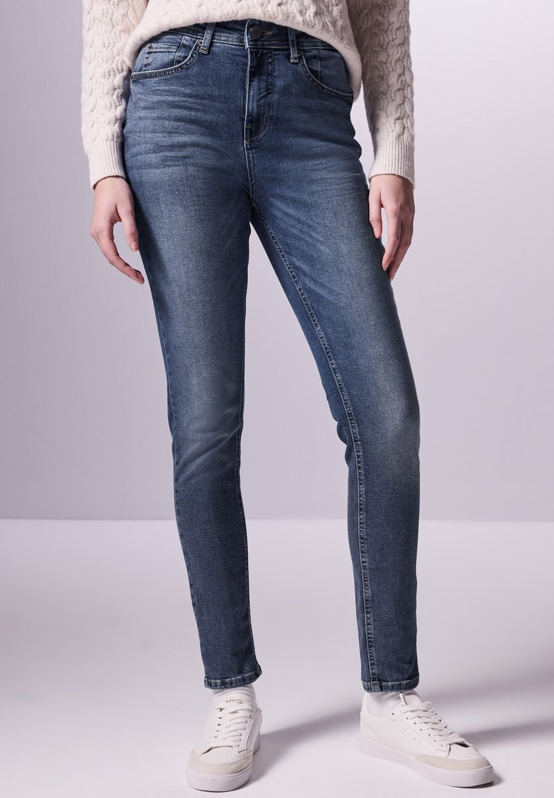 Jeans skinny en denim bleu foncé, avec une taille mi-haute, cinq poches et une texture lisse, associés à des baskets blanches.