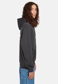 Sudadera gris oscuro con bolsillo canguro, capucha con cordón y puños de canalé. Textura suave y diseño minimalista. Combinada con jeans de color azul claro.