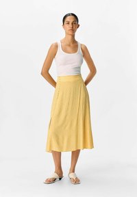 Jupe midi jaune avec un motif à pois, en tissu doux, fente sur le côté, portée avec un débardeur blanc et des sandales. Design simple et couleur vive.