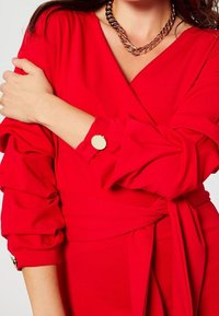 Rotes Wickelkleid mit voluminösen Ärmeln, taillierter Bund und goldfarbenem Knopfakzent. Glatter Stoff mit strukturiertem Design.