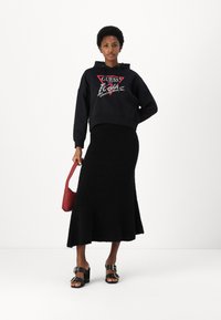 Felpa nera con logo "GUESS", abbinata a una gonna nera strutturata. La modella tiene una borsa rossa e indossa sandali neri con tacco e fibbie.