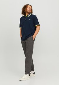 Marineblauw polo shirt met een groene en witte gestreepte kraag en mouwen, gecombineerd met grijze gestructureerde broek en witte sneakers met marineblauwe accenten.