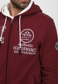 Felpa con zip color bordeaux, dotata di lacci bianchi, tasche frontali e branding stampato. Presenta un tessuto testurizzato e un rivestimento interno a contrasto.