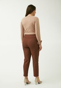 Lichtbeige langetermijn-top met knoopaccenten op de manchetten, gecombineerd met een strakke bruine broek en lichtbeige hoge hakken. Achteraanzicht.