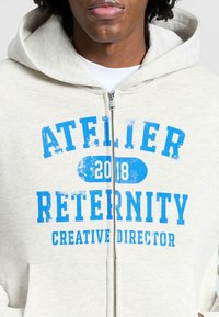 Młoda osoba w jasnoszarej bluzie z kapturem zapinanej na zamek, z niebieskim napisem "Atelier 2018 Reternity Creative Director" na piersi.