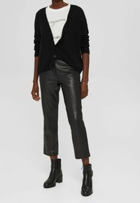 Schwarze, flauschige Strickjacke mit Knopfvorrichtung über einem weißen Grafik-T-Shirt. Kombiniert mit glänzenden schwarzen Lederhosen und Ankle Boots.