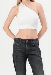 Donna che indossa un crop top bianco a spalla unica e jeans neri a vita alta, in piedi davanti a uno sfondo bianco semplice, con le mani dietro la schiena.