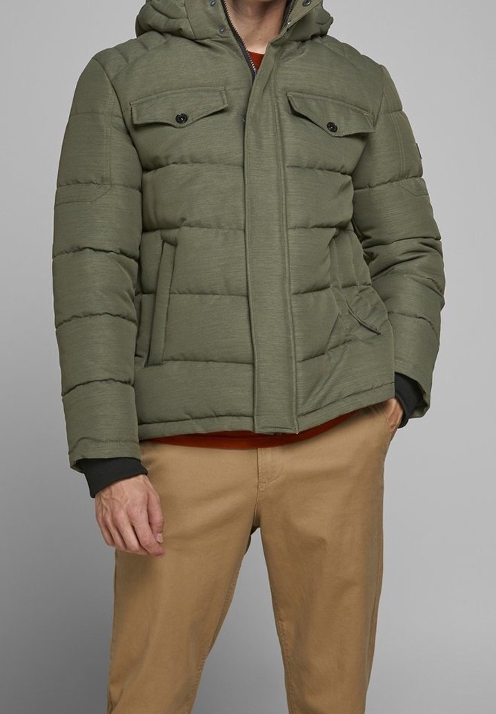 Doudoune verte à capuche, matelassée horizontalement, dotée de deux poches poitrine et d'une fermeture éclair frontale, portée avec un pantalon beige.