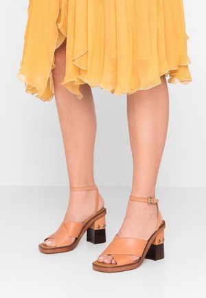 Jambes portant des sandales à talons carrés en cuir beige avec des brides autour des chevilles, associées à une jupe jaune fluide arrivant au genou.