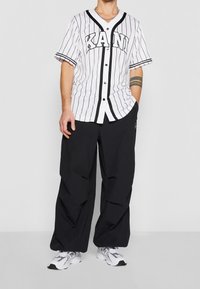 Vit basebolltröja med svarta vertikala ränder, "KANI" tryck, knappar fram, ihop med lösa svarta byxor och grå sneakers.