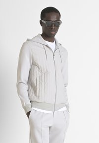 Felpa con cappuccio grigio chiaro trapuntata, con cerniera, polsini a costine e tasche laterali, indossata sopra una maglietta bianca a girocollo e pantaloni abbinati.
