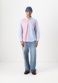 Camicia a maniche lunghe con pannelli rosa e azzurri chiari, maniche a righe e taschino sul petto. Indossata con jeans azzurri chiari e scarpe marroni.