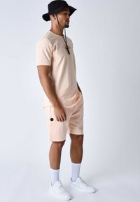 Maglietta rosa chiaro e pantaloni corti abbinati, con tasche laterali e un logo. Abbinati a sneakers bianche e un cappello a secchiello nero.