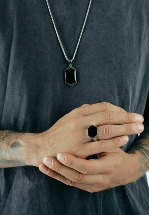 Black hexagonal pendant on a silver chain, paired with a matching hexagonal black ring. Worn on a bare hand against a dark shirt.