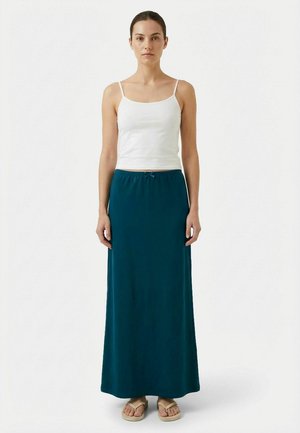 Žena stojaca čelom vpred, oblečená v bielom tielku bez rukávov, dlhej tmavomodrozelenej sukni a béžových žabkách na neutrálom pozadí.