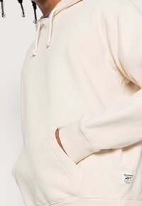 Krémová mikina s kapucí z měkké tkaniny, vybavená klokaní kapsou, žebrovanými manžetami, stahovací kapucí a malou visačkou s logem Reebok.