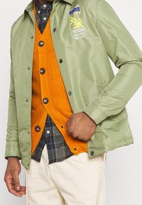 Grøn, let jakke med en blank finish, der har et gult grafisk patch. Underneath en orange tekstureret cardigan med mørke knapper.