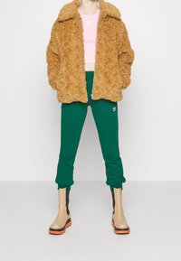 En fluffig brun zip-up jacka, en rosa crop top, gröna sweatpants och beige ankelstövlar med orange sulor. Outfiten kännetecknas av framträdande lager och textur.