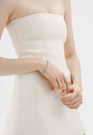 Persona con un vestido blanco sin tirantes con detalles de costuras verticales y una pulsera plateada, con las manos suavemente entrelazadas al frente.