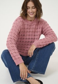 Maglione rosa lavorato a maglia con un design a motivo e maniche a costine. Abbinato a jeans in denim a gamba larga e scarpe nere.