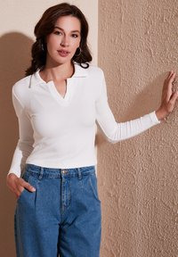 White ribbed long-sleeve top with a collar, paired with high-waisted blue denim pants featuring a front button and side pockets.