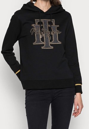 Person trägt einen schwarzen Hoodie mit goldbesetztem "TT"-Logo und dem kursiven Schriftzug "Tommy Hilfiger" auf der Brust, kombiniert mit schwarzen Hosen.
