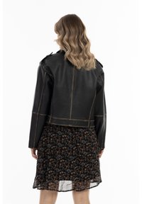 Zwarte leren cropped jas met zichtbare stiksels, gedragen over een jurk met bloemenprint en een gelaagde onderrand, die een gelaagde textuur laat zien.