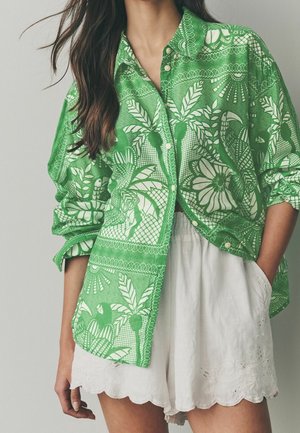 Femme portant une chemise verte à boutons avec un imprimé tropical blanc et un short blanc festonné avec des poches, debout avec une main dans la poche.