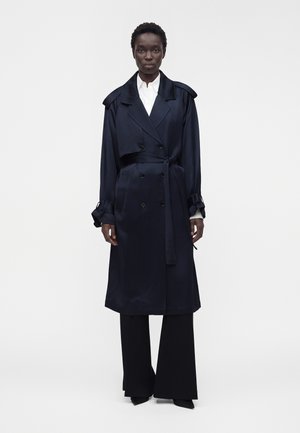 Vrouw die een lange, donkerblauwe, dubbelgeknoopte trenchcoat met riem draagt, wijdvallende zwarte broek, wit overhemd, staand tegen een effen achtergrond.