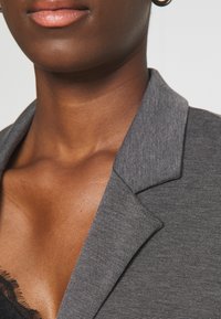 Blazer gris con una tela texturada, solapas afiladas y un diseño ajustado, combinado con una blusa de encaje negra visible por debajo.