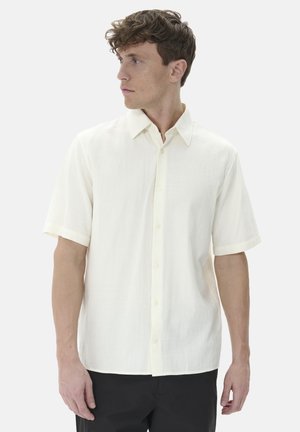 Camisa de manga corta, abotonada, en tela de color beige claro, con un collar clásico y una textura sutil, combinada con pantalones negros.