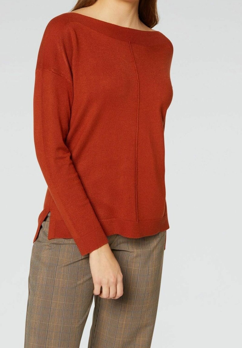 Pull de couleur rouille avec un col large et des manches longues, présentant une finition texturée et des fentes latérales, associé à un pantalon rayé.