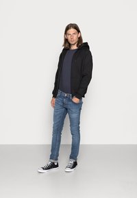 Hanorac cu fermoar negru, cămașă bleumarin, blugi slim-fit de culoare albastru deschis și adidași negri cu gât înalt. Design minimalist, cu manșete striate și buzunare.