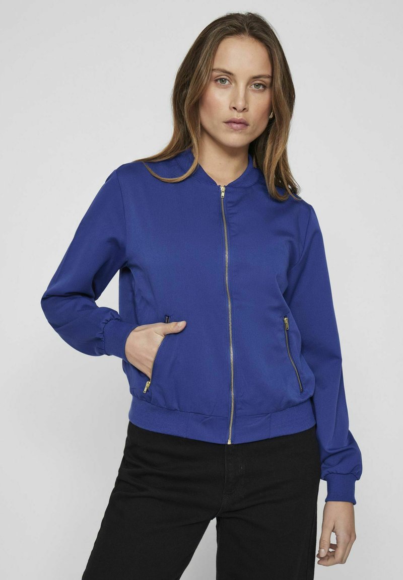 Niebieska kurtka bomberka z zamkiem z przodu, elastycznymi mankietami i dwiema bocznymi kieszeniami. Gładki materiał o jednolitym kolorze i złotych akcentach zamka.