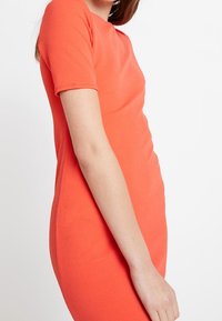 Kortärmad, figursydd klänning i ljus korall, tillverkad av texturerat tyg. Enkel rund halsringning och slät silhuett.